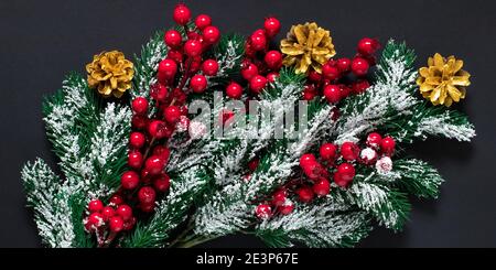 Weihnachtsbaumschmuck auf dunklem Hintergrund. Grüne Tannenzweige mit Schnee, goldene Tannenzapfen und rote Stechbeeren. Neujahrsstimmung, Winterfest Stockfoto