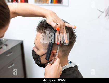 Männlicher Klient, der während einer Coronavirus-Pandemie einen Haarschnitt trägt. Nahaufnahme der Hände eines Barbiers, die mit einer Haarschere arbeiten. Stockfoto