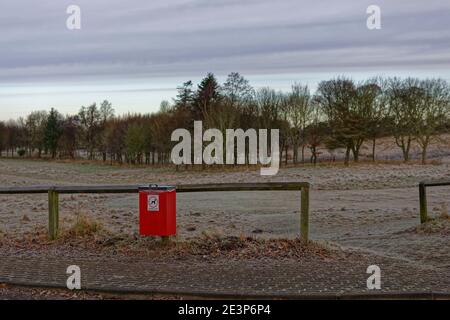 Eine rote Hundehüte mit Schild neben einem hölzernen Zaun Geländer, auf einem Bürgersteig bei einem Frosted Fairway des Letham Grange Golf Course. Stockfoto