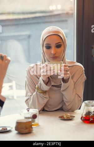 Trinken, reden. Schöne arabische Frau treffen im Café oder Restaurant mit Freund, Zeit mit einer Tasse Tee zu verbringen. Muslimischer Lebensstil. Stilvolles und glückliches Modell mit Make-up in komfortabler Innenausstattung. Stockfoto