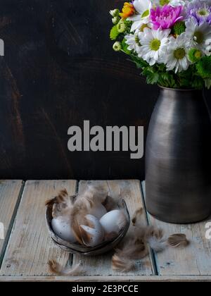 Vase mit buntem Blumenstrauß und dekorativen Eiern mit Federn auf Holztisch. Speicherplatz kopieren. Dunkel und launisch. Stockfoto