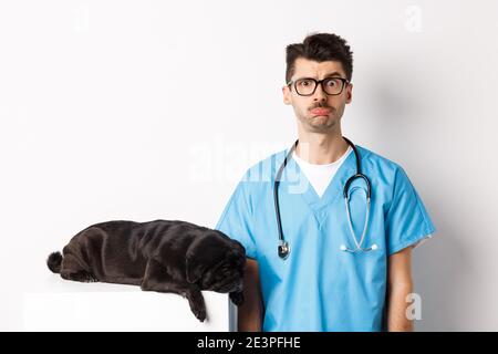 Müde schwarzen Hund Mops liegen in der Nähe von schönen männlichen Arzt in der Tierklinik, Tierarzt starrte verwirrt auf Kamera, über weißem Hintergrund stehen Stockfoto