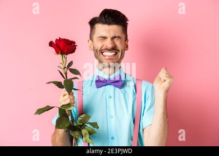 Romantik und Valentinstag Konzept. Hoffnungsvoller Mann, der sich vor Date aufgeregt fühlt, auf Liebhaber mit roter Rose wartet und betet, über Rosa steht Stockfoto