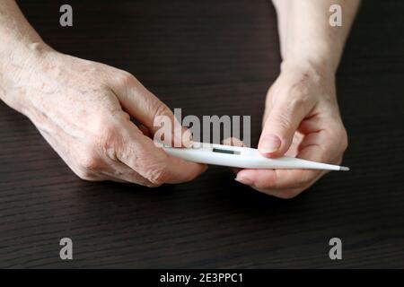 Ältere Frau misst Körpertemperatur, Konzept von Fieber, Coronavirus-Symptome. Digitalthermometer in faltigen weiblichen Händen aus der Nähe Stockfoto