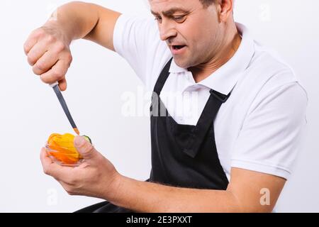 Vergrößertes Foto eines männlichen Kochs in einer schwarzen Schürze mit einer Schüssel Gemüse und einem Messer auf weißem Hintergrund. Stockfoto