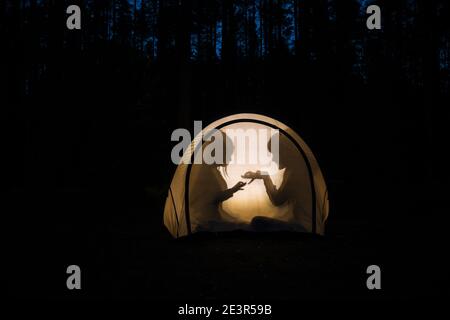 Silhouetten von Kindern, die nachts im Zelt spielen, machen Schattenpuppen mit Taschenlampe und genießen Sommerferien Stockfoto