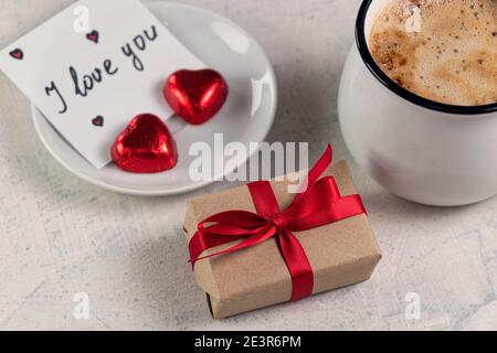 Ein Geschenk mit einem roten Band, einer Tasse Kaffee, herzförmiger Schokolade und einer Note mit dem Wort Ich liebe dich. Valentinstag Frühstück. Valentinstag-Konzept. Weichfokus Stockfoto