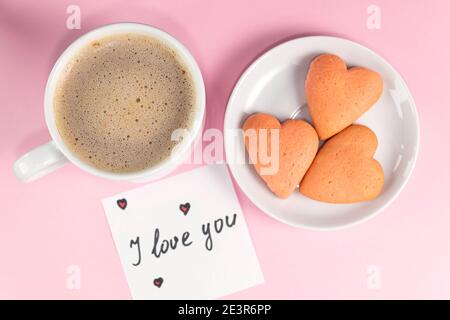 Tasse Kaffee, hausgemachte herzförmige Kekse und eine Notiz mit den Worten I love you auf pastellrosa Farbe. Valentinstag-Frühstück. Valentinstag-Konzept. Weichfokus Stockfoto