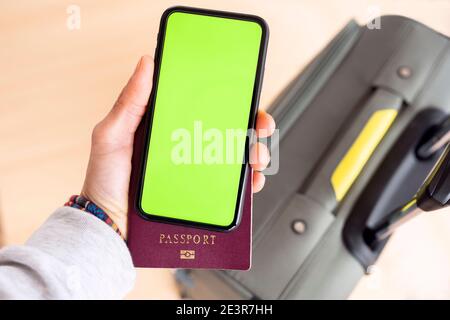 (Selektiver Fokus) Ansicht von oben einer menschlichen Hand mit einem Pass und einem Smartphone mit einem grünen Bildschirm und einem Unschärfegepäck im Hintergrund. Stockfoto