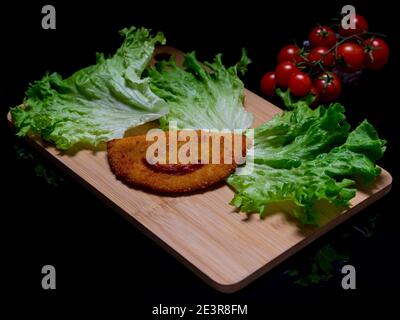 Ein schmackhafter gebratener Sofficino mit einem Lächeln, der mit Ketchup, Salatblättern und Kirschtomaten auf Holzschneidebrett und schwarzem reflektierendem Hintergrund hergestellt wird Stockfoto