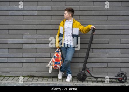 Teenager mit Rucksack und Elektroroller auf Backstein Wand Hintergrund. Modernes Schuljunge Konzept Stockfoto