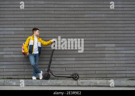 Teenager mit Rucksack und Elektroroller auf Backstein Wand Hintergrund. Modernes Schuljunge Konzept Stockfoto