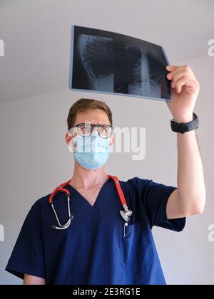 Ein hübscher Arzt, der Peeling, Stethoskop und Gesichtsmaske trug und während der sars COV 2 Pandemie Covid 19 auf eine Röntgenaufnahme blickte Stockfoto