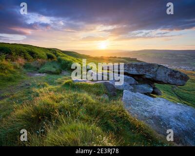 Die untergehende Sonne fällt unter eine Wolkenbank und taucht die Landschaft rund um den Pancake Rock am Rande des Ilkley Moor in goldenes Licht. Stockfoto