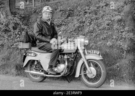 Vintage Schwarz-Weiß-Fotografie eines Mannes mit Sturzhelm und Brille oben, sitzend auf einem Triumph Twenty One Motorrad, Kennzeichen 753 BYW. Das Motorrad ist auf seinem Ständer geparkt und läuft nicht. Stockfoto