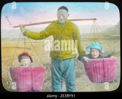 Mann, der zwei kleine Kinder in den Körben eines Joches trägt, China, ca. 1917-1923 (IMP-YDS-RG224-OV1-0000-0030). Stockfoto