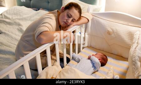 Junge müde und erschöpft Mutter eingeschlafen, während Schaukeln Krippe ihres neugeborenen Babys in der Nacht. Konzept der schlaflosen Nächte und Elterndepression Stockfoto