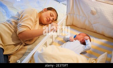 Junge müde Mutter schlief ein, während sie ihren kleinen Sohn in der Krippe in der späten Nacht schaukelte. Mutterschaft und schlaflose Nächte. Stockfoto