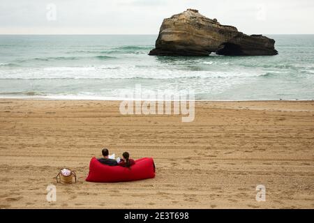 Paar entspannt auf einem roten aufblasbaren Luftsofa am Strand von Biarritz mit Blick auf einen natürlichen Meeresfelsenbogen im Atlantischen Ozean an einem bewölkten Frühlingstag, Frankreich Stockfoto