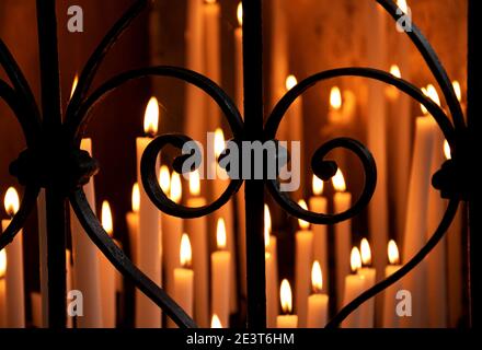 Goldene Kerzen leuchten im dunklen Tempel durch Schmiedezaun in Herzform gesehen. Religiöser Hintergrund. Selektiver Fokus auf den Zaun. Verschwommenes Licht. Stockfoto