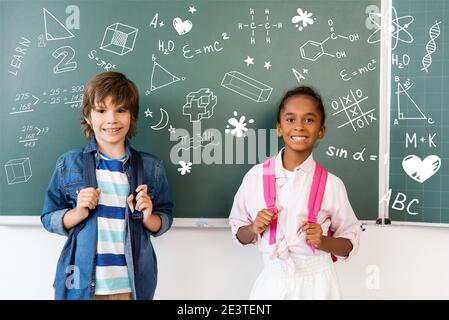 Schuljunge mit Rucksack in der Nähe afroamerikanischen Freund und Kreidetafel stehen Mit Illustration im Klassenzimmer Stockfoto