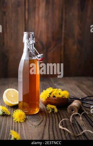 Hausgemachter gesunder Löwenzahn-Sirup in einer Glasflasche, dekoriert mit frischen Blumen auf rustikalem Holzhintergrund mit Kopierraum Stockfoto