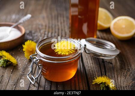 Hausgemachter gesunder Löwenzahn-Sirup in einer Glasflasche, dekoriert mit frischen Blumen auf rustikalem Holzhintergrund Stockfoto
