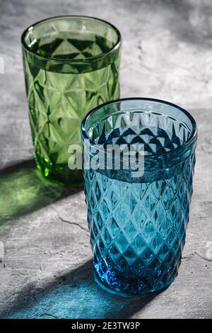 Zwei geometrische Glas Tasse mit Wasser in blauen und grünen Farben mit bunten Schatten Lichtstrahlen auf Stein Beton Hintergrund, Winkel Ansicht Stockfoto