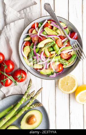 Gesundes veganes Gericht, saftiger Sommersalat mit blanchierten Spargel, Kirschtomaten, Avocado-Scheiben und roter Zwiebel, mit Pfeffer bestreut und mit Nieselteich beträufelt Stockfoto