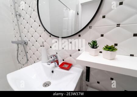 Moderne weiße und schwarze WC-Innenausstattung. Weißes Waschbecken mit Wasserhahn und Toilettenartikeln, darüber großer runder Spiegel an der Wand der Keramikfliesen Stockfoto