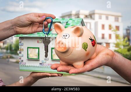 Immobilienmakler geben flache Schlüssel zu einem neuen Eigentümer, der letzte Geld sparen von Sparschwein auf verschwommenem Hintergrund zahlt Stockfoto