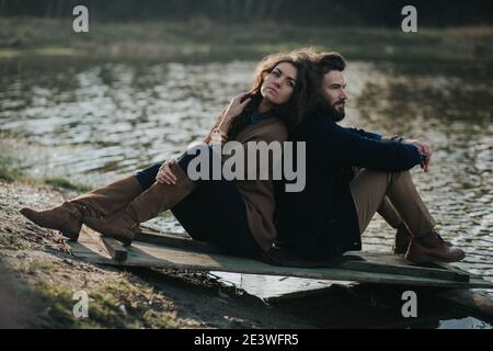 Zwei kaukasische Liebhaber sitzen auf dem Pier am See. Das junge Paar umarmt sich am Herbsttag im Freien. Ein bärtiger Mann und eine lockige Frau in der Liebe. Valentinstag Stockfoto
