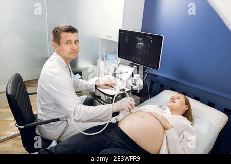 Horizontales Bild von schönen jungen männlichen Arzt, Blick auf die Kamera, während der Durchführung Ultraschall-Diagnoseverfahren auf schwangere Frau in der modernen Klinik Stockfoto