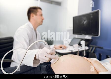 Nahaufnahme eines männlichen Arztes, der Ultraschall-Sonogramm-Verfahren bei nicht erkennbarer schwangerer Frau durchführt. Konzentrieren Sie sich auf den Ultraschallaufnehmer am Stockfoto