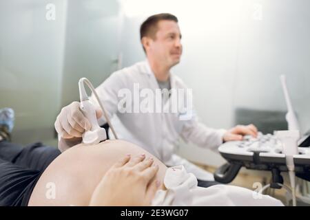 Beschnitten Nahaufnahme eines Bauches einer schwangeren Frau mit Ultraschall-Scanning in der medizinischen Klinik. Männlich gut aussehend reifen Arzt machen Ultraschall Stockfoto