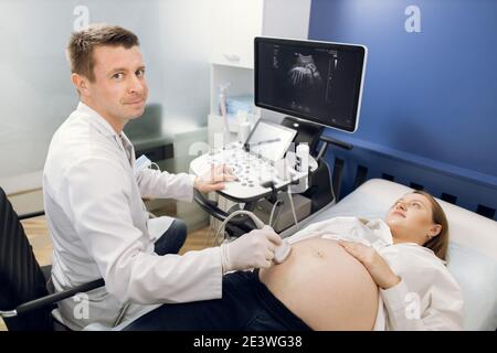 Lächelnder Mann Geburtshelfer Untersuchung schwangeren Bauch durch Ultraschall-Scan. Glückliche schwangere Dame besucht ihren Arzt in der modernen Entbindungszentrum, tun Stockfoto