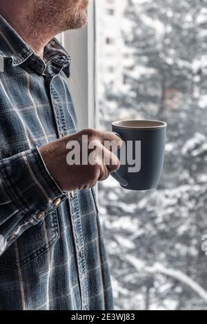 Ein blauer Becher mit Kaffee in den Händen eines Mannes vor dem Hintergrund eines Fensters mit schneebedeckten Bäumen an einem Wintermorgen. Wärmende Getränk im Winter. Stockfoto