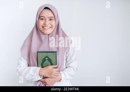 Junge asiatische Islam Frau trägt Kopftuch hält heiligen al quran in der Hand mit Lächeln und glückliches Gesicht. Indonesierin auf grauem Hintergrund. Stockfoto