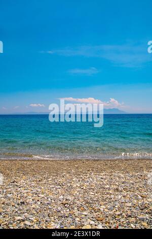 Schöner vertikaler Meerblick vom Ufer bis zu tropisch klarem türkisfarbenem Wasser. Insel am Horizont und einige weiße Wolken am blauen Himmel. Bunte Kieselsteine Stockfoto