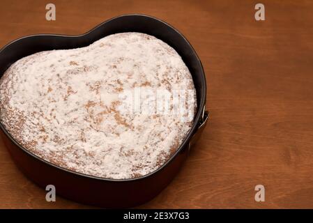 Herzförmiger Kuchen, der in einer Springform mit Puderzucker bestäubt ist Auf einem Holztisch Stockfoto