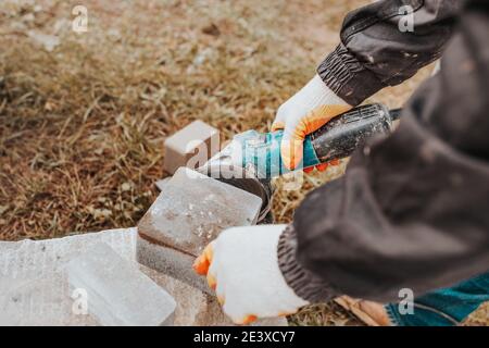 Ein Maurer schneidet einen Betonblock aus Pflasterplatten Mit einem Schleifer - Einstellung der Fliesen beim Verlegen Stockfoto
