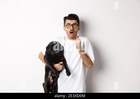 Erschrockener Hipster umarmt seine niedlichen schwarzen Welpen und starrte auf Kamera erschrocken. Hundebesitzer schaut schockiert, hält Mops, steht auf weißem Hintergrund Stockfoto