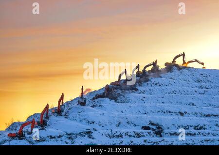 Silhouetten von vielen Baggern, die auf einem riesigen Berg arbeiten Eine Müllhalde vor dem Hintergrund eines orangefarbenen Sonnenaufgangs Oder Sonnenuntergang Himmel im Winter Stockfoto