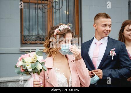 Die Braut in einer medizinischen Maske mit dem Bräutigam in der Nähe des Standesamtes. Hochzeit während der covid-19 Epidemie, Schutz vor dem Virus in öffentlichen Räumen. Stockfoto