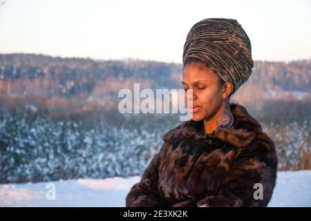 Schönes Mädchen mit Pelz Mantel und Turban genießen Sonnenuntergang oder sonnenaufgang auf dem Schnee im Winter mit Waldbäumen und Sonnenuntergang im Hintergrund Stockfoto