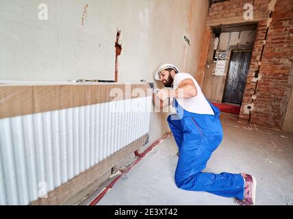 Klempner, trägt blaue Overalls, weißer Helm, Installation Wasserkühler, Arbeit mit Schraubenschlüssel. Wartung Reparaturarbeiten Renovierung in der Wohnung. Weitwinkel. Bau- und Hausrenovierungskonzept. Stockfoto