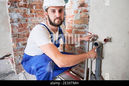 Schöner männlicher Arbeiter in Schutzhelm Blick auf Kamera, während die Anwendung von Silikonfett auf Rohr. Mann Klempner in der Arbeit Overalls Schmierrohr bei der Installation von Wassersystem in der Wohnung. Stockfoto