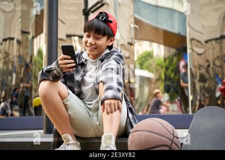 Teenager asiatische Kind mit Basketball und Skateboard auf Bordstein sitzen Von Straße auf Handy schauen Stockfoto