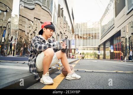 Teenager asiatische Kind mit Basketball und Skateboard auf Bordstein sitzen Von Straße auf Handy schauen Stockfoto