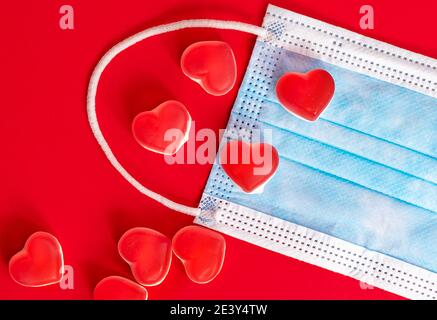 Blaue medizinische Maske mit Herzen auf rotem Hintergrund. Valentinstag 2021. Copyspace Stockfoto